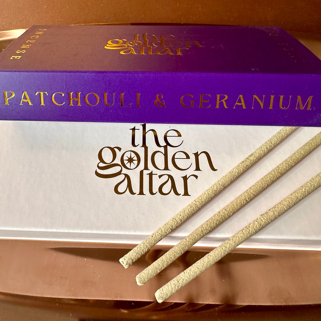 The Golden Altar Patchouli & Geranium Räucherstäbchen – sonniger Duft mit goldenem Schriftzug. Im Hintergrund ist die lila Verpackung, ausgepackte vegane Räucherstäbchen liegen auf der weißen Box mit gold geprägtem Namen.